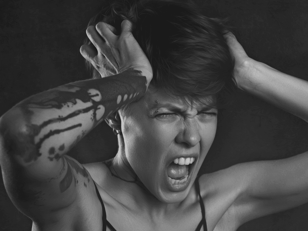 A black and white portrait of a person with tattooed arms gripping their head and screaming, eyes shut, against a dark background