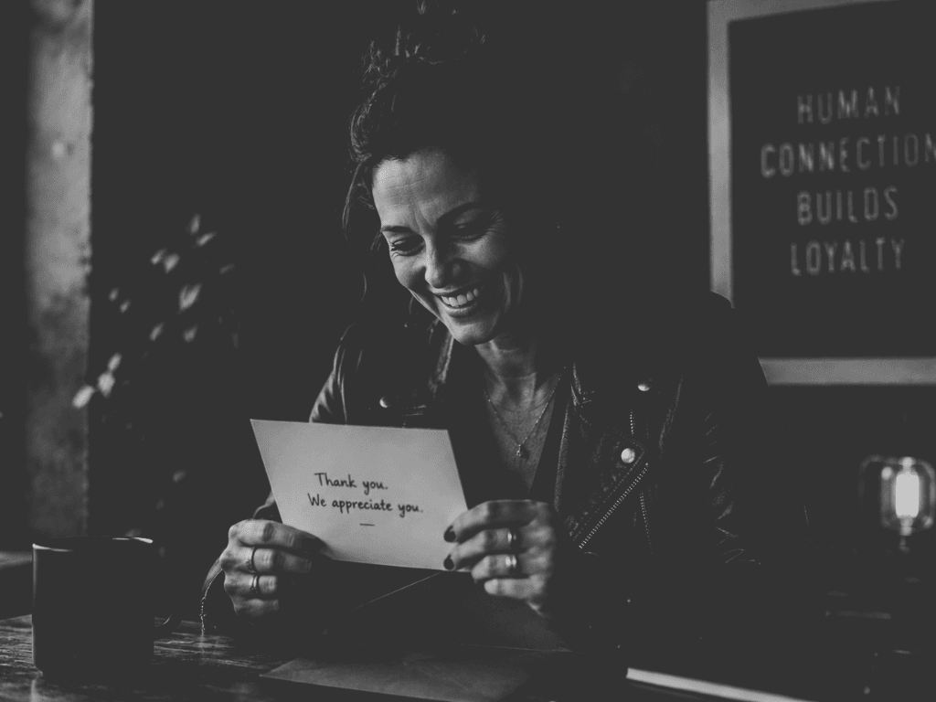 A smiling woman reading a handwritten thank-you note in a cafe, with a sign behind her reading Human Connection Builds Loyalty