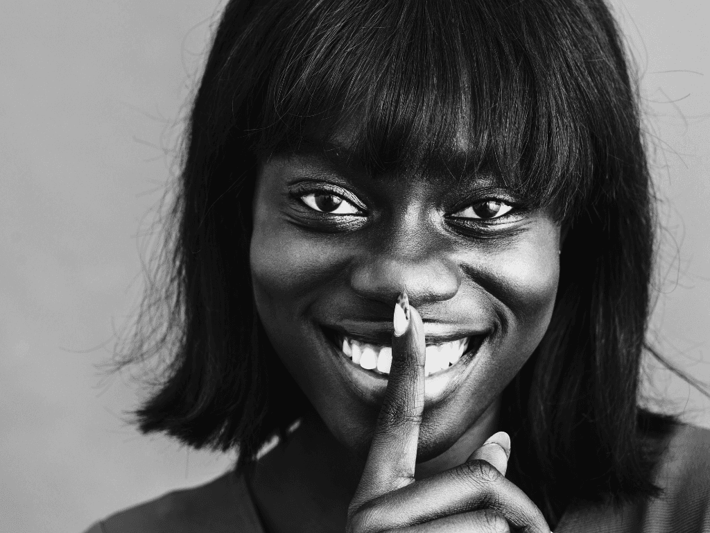 A woman smiling directly at the camera with her finger pressed to her lips in a 'shhh' gesture, shot in black and white