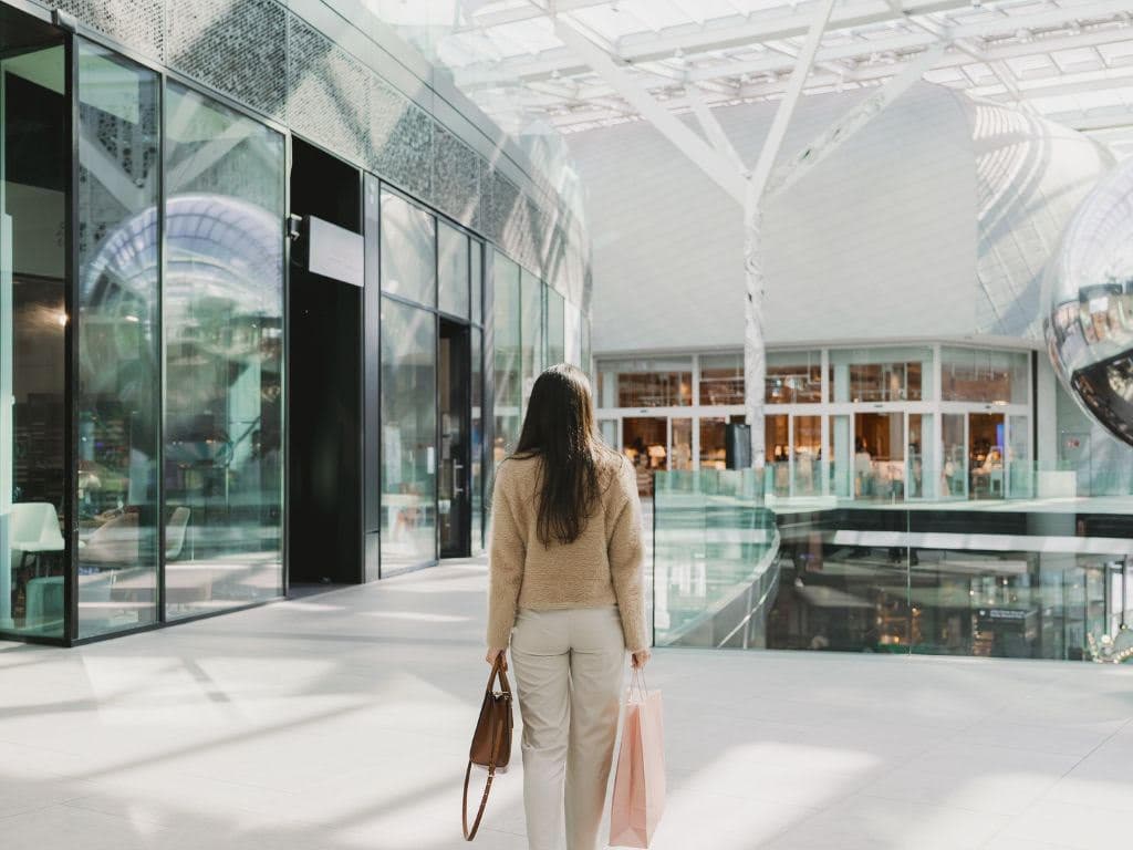 A woman with dark hair wearing a cream sweater walking through a modern glass shopping centre carrying a brown leather handbag and a pink shopping bag