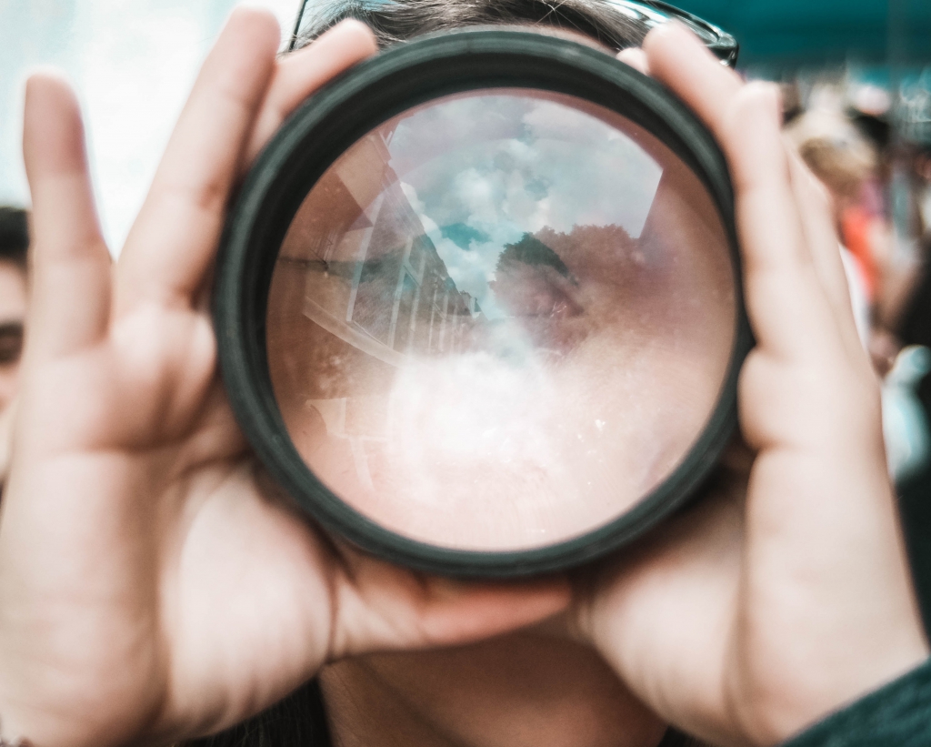 A person holding a camera lens up to their face, city reflected inside the glass