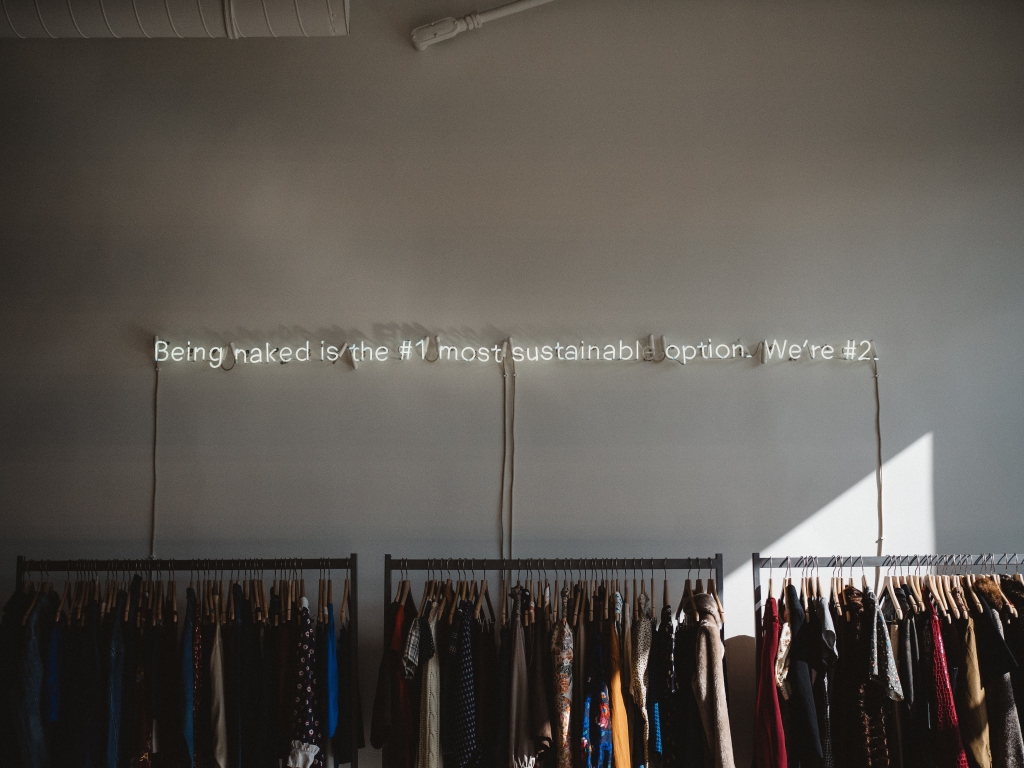 Clothing rails beneath a neon sign reading 'Being naked is the #1 most sustainable option. We're #2.'