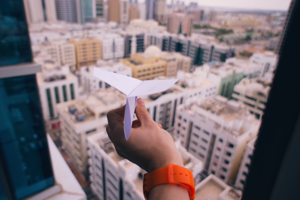 A hand holding a paper plane out of a high-rise window over a city skyline